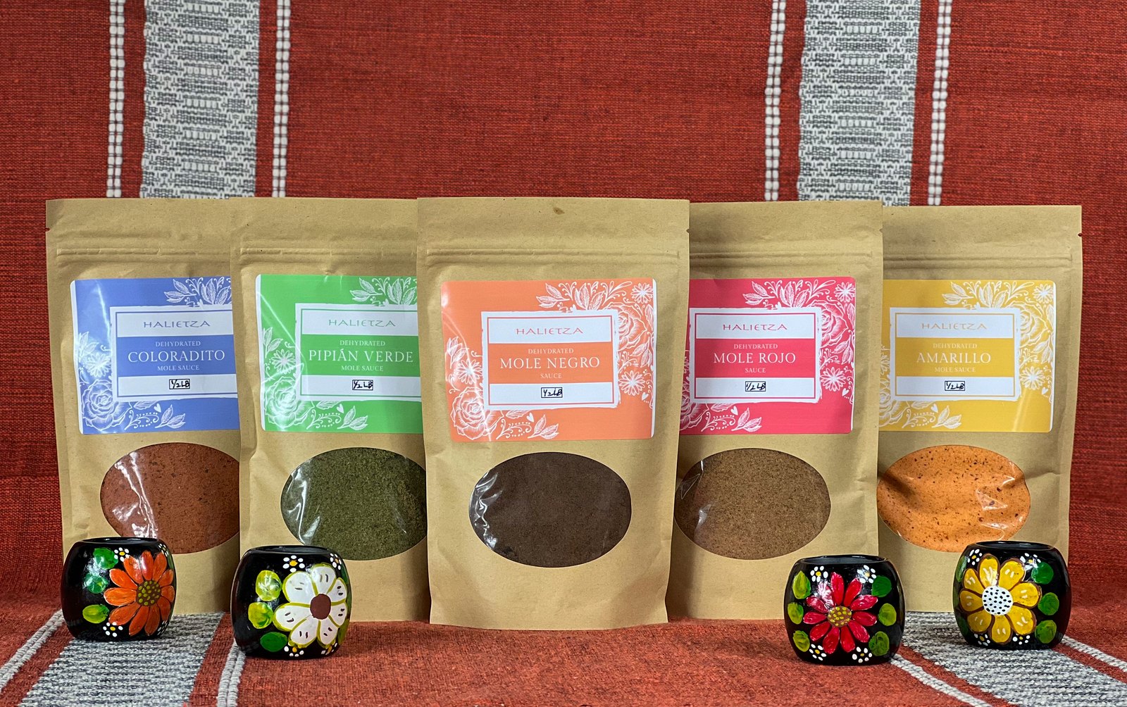 The first five Halietza pouches, Coloradito, Pipián Verde, Mole Negro, Mole Rojo and Amarillo, in kraft bags with a window, on a red woven cloth