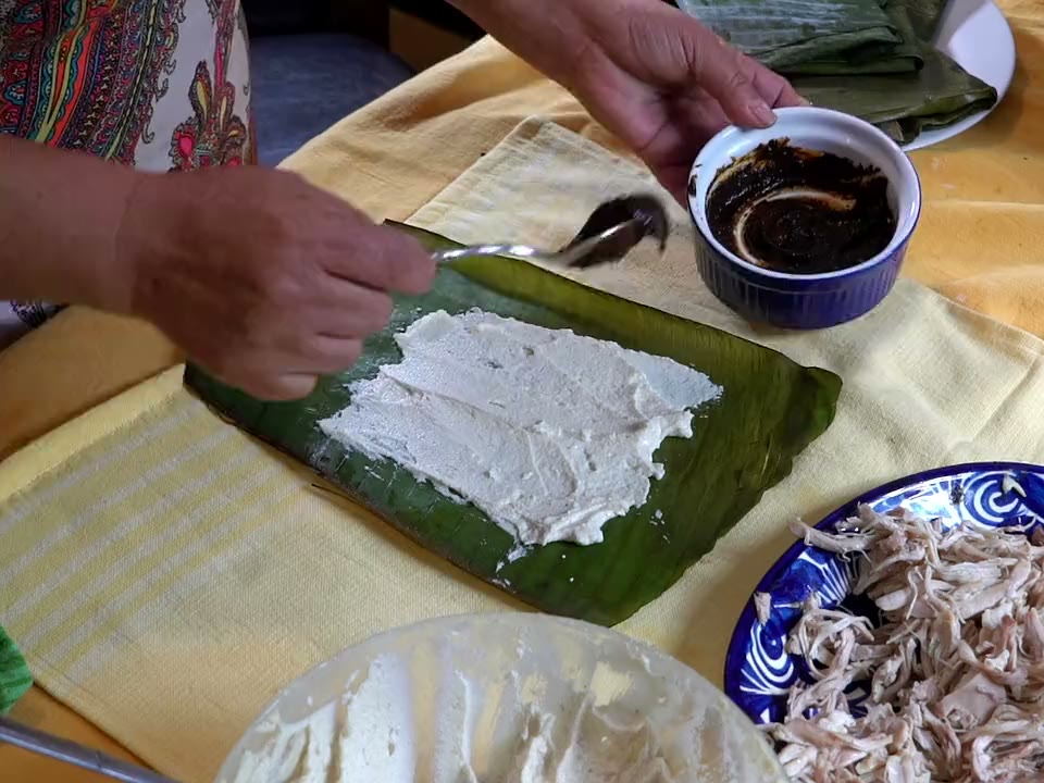 Elisa Beristain's hands spooning mole negro onto the masa of a banana leaf tamal, beside a plate of shredded chicken